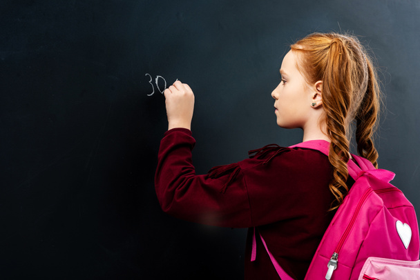 schoolgirl with pink backpack writing on blackboard with chalk - Foto, afbeelding