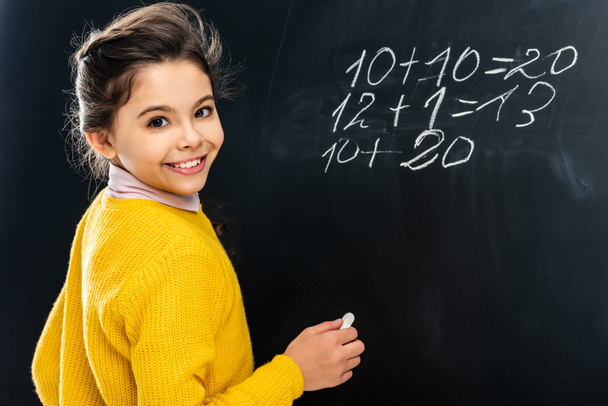 cheerful schoolgirl in yellow holding chalk and looking at camera in front of blackboard - Photo, Image