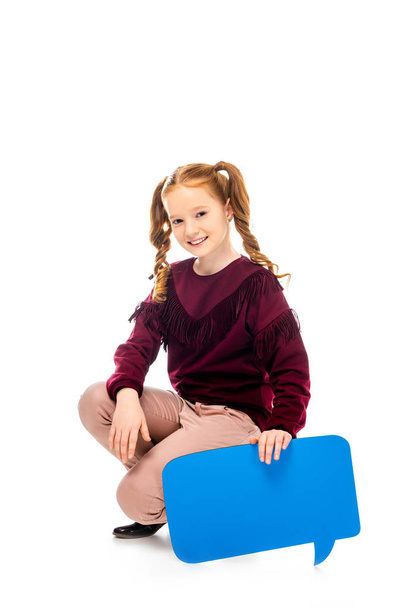 smiling schoolgirl sitting on hunkers, holding speech bubble and looking at camera isolated on white - Fotoğraf, Görsel
