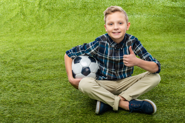 smiling boy holding soccer ball, looking at camera and showing thumb up on lawn - Foto, Imagen