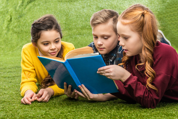 cute preteen schoolkids lying on green lawn and reading book - Photo, Image