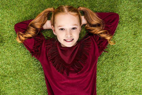 top view of smiling schoolgirl with hands on head looking at camera and lying on lawn - Foto, Imagen