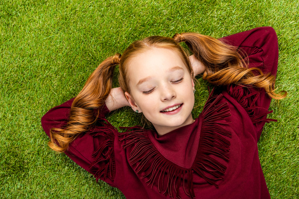 top view of cute schoolgirl lying on lawn with closed eyes - Foto, Imagen