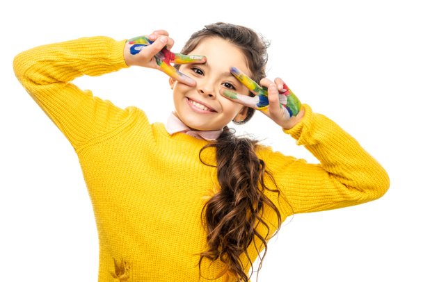 aranyos iskoláslány a sárga pulóver látszó-on fényképezőgép, és béke jeleit, kézzel festett színes festékek elszigetelt fehér - Fotó, kép