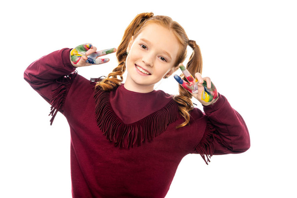 smiling schoolgirl looking at camera and showing peace signs with hands painted in colorful paints isolated on white - Photo, Image