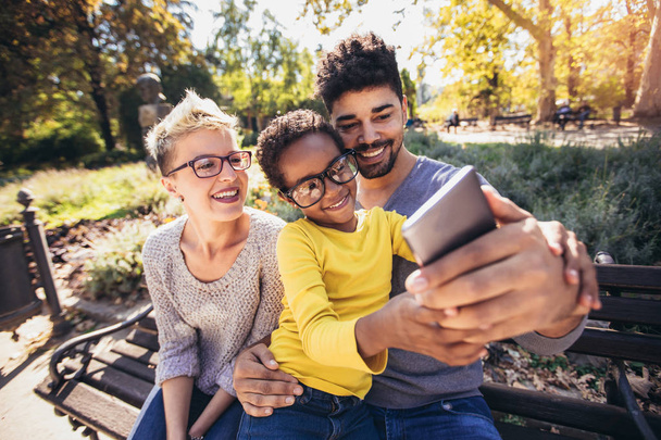 Happy young mixed race couple spending time with their daughter using smart phone in public park - Foto, Imagem