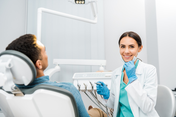 alegre dentista sosteniendo taladro y sonriendo cerca africano americano paciente
  - Foto, Imagen