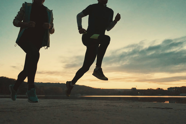 Mladý pár běžců nohy běží za úsvitu s jezerem a obloze v pozadí. Sportovně založení lidé trénink venku. Dolů, úhel záběru. - Fotografie, Obrázek