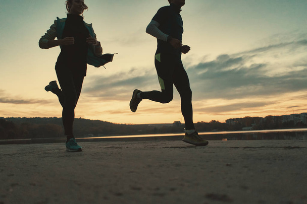 Ein motiviertes Läuferpaar machte sich bei Sonnenaufgang auf den Weg zum See. junge Männer und Frauen in Sportkleidung, die zusammen laufen. Sportler trainieren im Freien - Foto, Bild