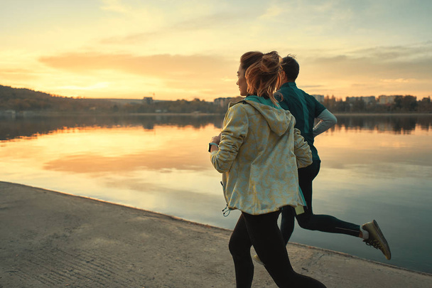 Un par de corredores motivados a correr por el lago al amanecer. Joven hombre y mujer en ropa deportiva, corriendo juntos. Formación de deportistas al aire libre
 - Foto, Imagen