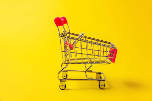 Small supermarket grocery push cart for shopping toy with wheels isolated on yellow colourful trendy modern fashion background. Sale buy mall market shop consumer concept. Copy space - Photo, Image