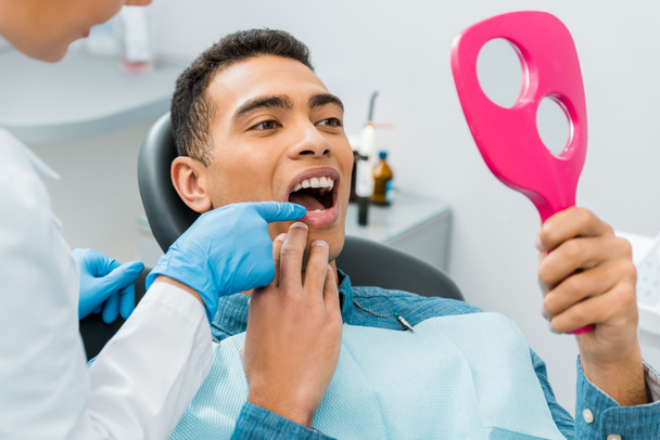 kobiece dentysta dotykając zębów przystojny mężczyzna african american, patrząc w lustro  - Zdjęcie, obraz