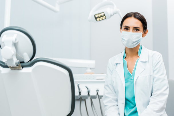 female dentist in white coat and mask near dental chair  - Foto, imagen
