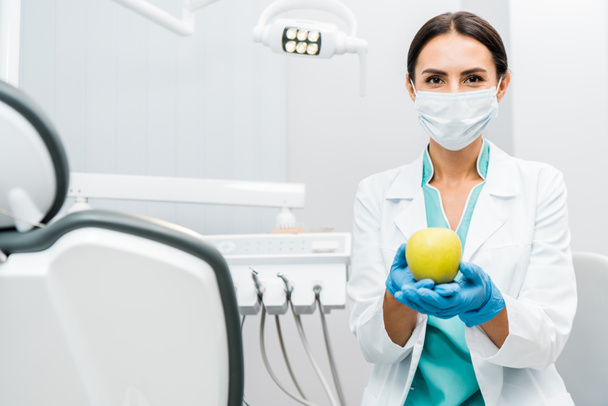 female dentist in white coat and mask holding apple  - Photo, Image