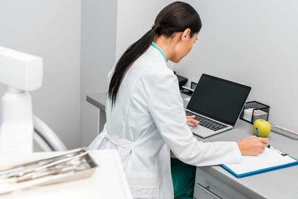 medico femminile utilizzando computer portatile con schermo bianco e prendere appunti
 - Foto, immagini