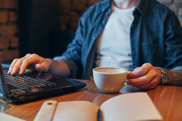 Hombre joven bebiendo café en la cafetería de la ciudad durante el almuerzo y trabajando en el ordenador portátil. De cerca.
 - Foto, imagen