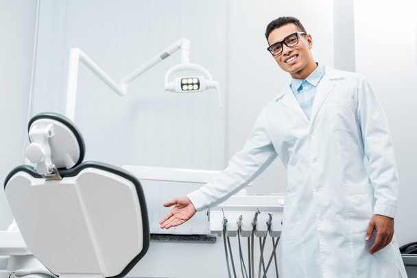 χαμογελώντας Αφρικανικός Αμερικανός γιατρός δείχνει καρέκλα σε οδοντιατρείο  - Φωτογραφία, εικόνα