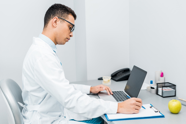 handsome african american doctor making notes and using laptop  - Foto, Bild