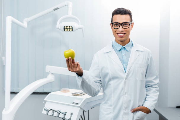smiling african american dentist with hand in pocket holding apple - Fotoğraf, Görsel