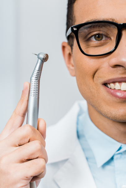 κοντινό πλάνο της χαμογελώντας Αφρικανικός Αμερικανός οδοντίατρος σε ποτήρια κρατώντας τρυπάνι  - Φωτογραφία, εικόνα