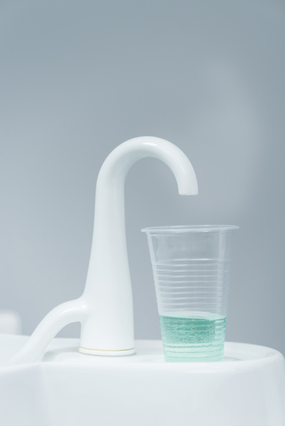 Close up van plastic beker met vloeistof in de buurt van witte kraan in tandheelkundige kliniek  - Foto, afbeelding