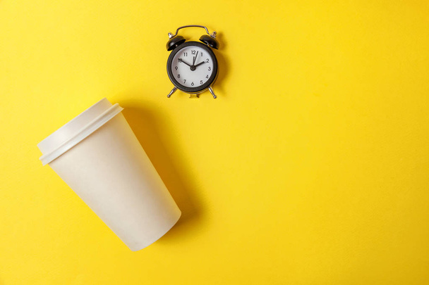 単にフラットなレイアウト デザイン紙のコーヒー カップと黄色のカラフルなトレンディな背景の目覚まし時計。テイクアウトのドリンクと朝食飲料。おはよう目を覚ます目を覚まし概念。トップ ビュー コピー スペース - 写真・画像