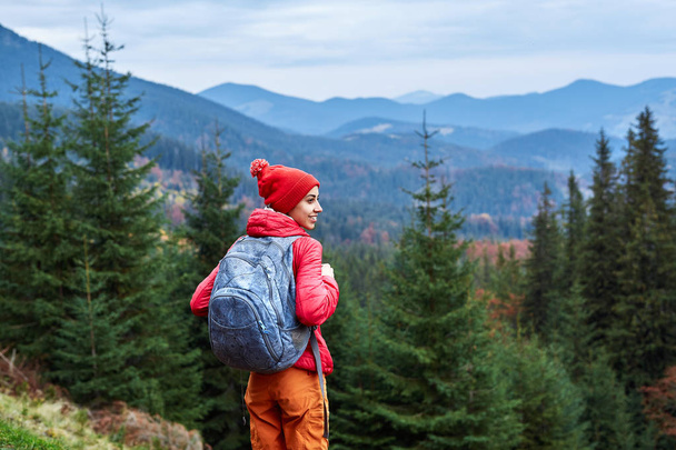 woman hiker with backpack, wearing in red jacket and orange pants, standing on the mountains background - Foto, Imagen