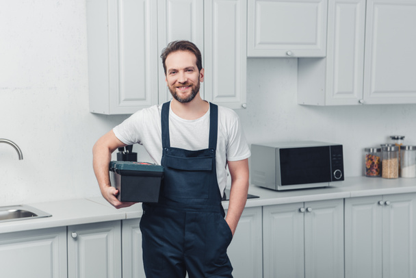 sonriente barbudo reparador en el trabajo en general la celebración de la caja de herramientas en la cocina en casa
 - Foto, Imagen