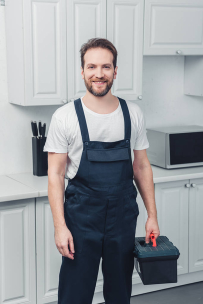 adult smiling bearded repairman in working overall holding toolbox in kitchen at home - Foto, Imagem
