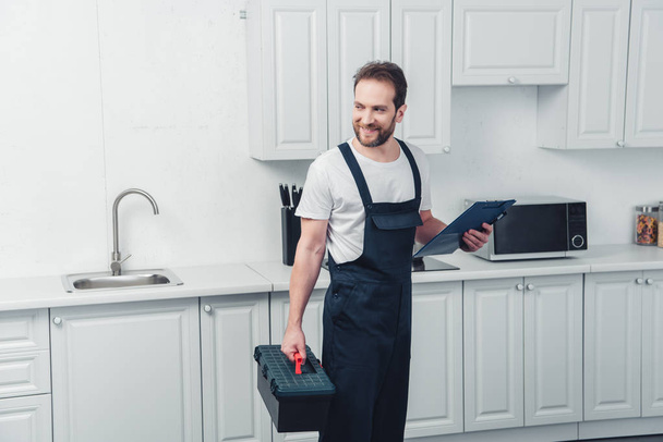 cheerful adult repairman in working overall holding toolbox and clipboard in kitchen at home - Valokuva, kuva