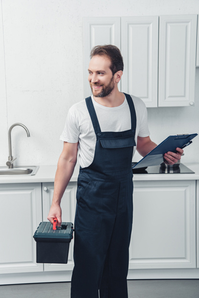 happy adult repairman in working overall holding toolbox and clipboard in kitchen at home - 写真・画像