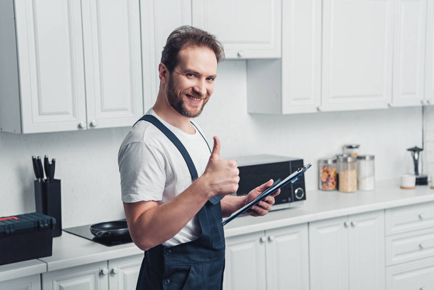 smiling handsome adult bearded repairman holding clipboard and doing thumb up gesture in kitchen  - Foto, Imagem