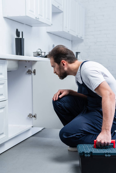 focused bearded repairman with toolbox checking sink in kitchen - Foto, Imagem