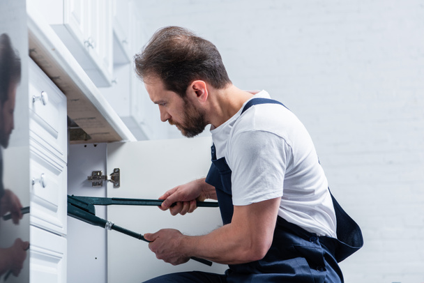 vista lateral del reparador barbudo adulto en el fregadero de fijación general de trabajo con un par de pinzas en la cocina
  - Foto, Imagen