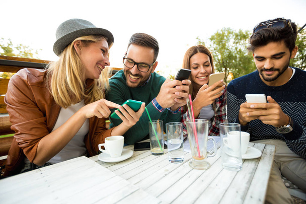 Group of four friends having fun a coffee together. Two women and two men at cafe talking laughing and enjoying their time. Using phone  - Zdjęcie, obraz