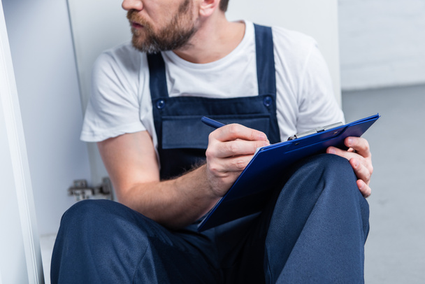 vista parcial del artesano adulto escribiendo en portapapeles mientras está sentado en el suelo en la cocina
 - Foto, Imagen