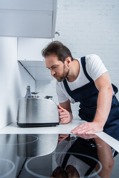 concentrated bearded craftsman in working overall fixing toaster in kitchen - Photo, Image