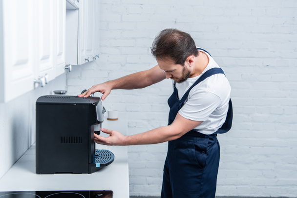 side view of handyman in working overall repairing coffee machine in kitchen - Photo, Image