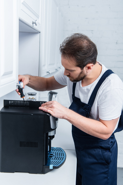 adult handyman in working overall repairing coffee machine by screwdriver in kitchen - Foto, Imagen