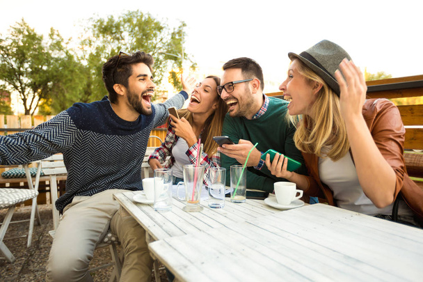 Neljän ystävän ryhmä pitää hauskaa kahvilla. Kaksi naista ja kaksi miestä kahvilassa nauramassa ja nauttimassa ajastaan. Puhelimen käyttö
  - Valokuva, kuva