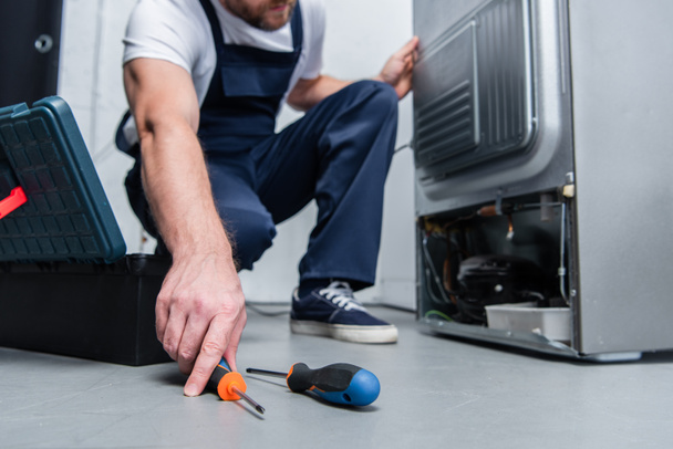 cropped shot of repairman in working overall taking screwdriver from floor near broken refrigerator in kitchen - Fotografie, Obrázek