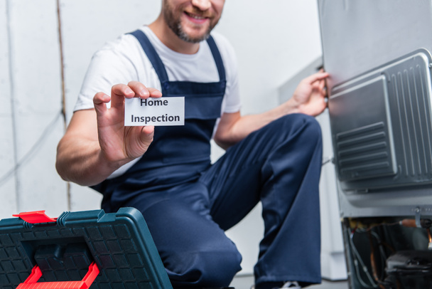 vista parcial del artesano adulto que muestra la tarjeta con la inspección de la casa de letras mientras está sentado cerca del refrigerador roto
  - Foto, Imagen