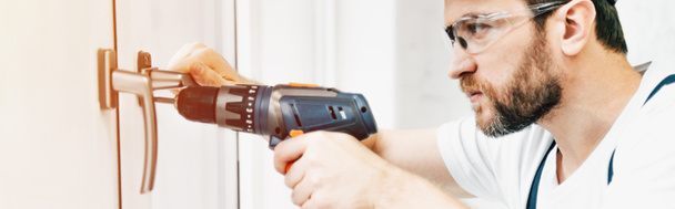 panoramic view of focused male handyman in goggle repairing window handle with electric drill  - Photo, Image