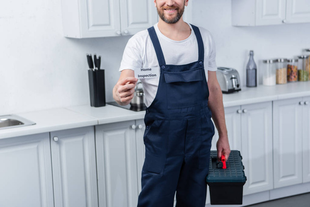 partial view of male handyman holding toolbox and showing card with lettering home inspection - Fotografie, Obrázek