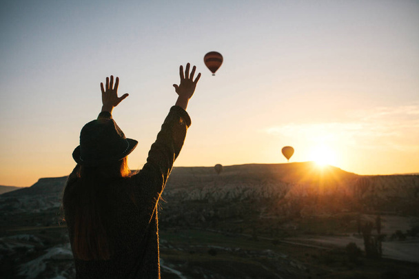 Ένα κορίτσι τουρίστας σε ένα καπέλο θαυμάζει αερόστατα θερμού αέρα πετώντας στον ουρανό πάνω από την Καππαδοκία, Τουρκία. Εντυπωσιακό θέαμα. - Φωτογραφία, εικόνα