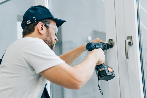 handyman in goggles fixing window handle by electric drill  - Φωτογραφία, εικόνα