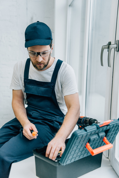 adult male repairman taking screwdriver from toolbox while sitting on windowsill - Φωτογραφία, εικόνα