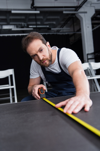 focused male craftsman in working overall making measurements of floor by measuring tape - Foto, Imagem
