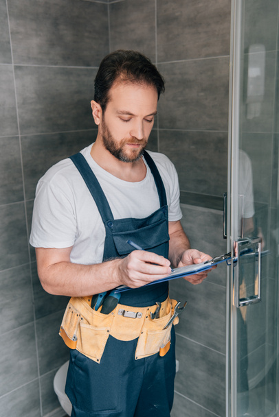 bearded male plumber making notes in clipboard while checking shower in bathroom - Fotografie, Obrázek