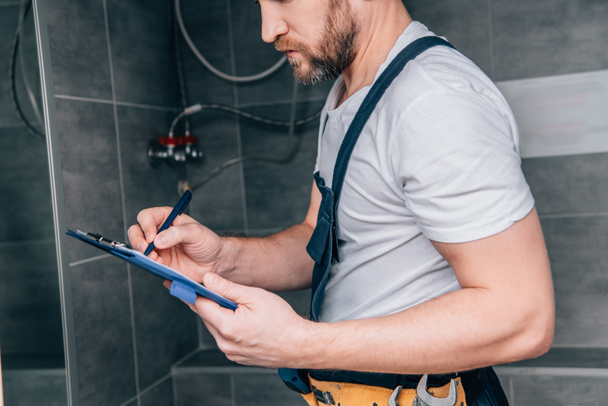 partial view of plumber writing in clipboard and checking electric boiler in bathroom  - Foto, Imagem